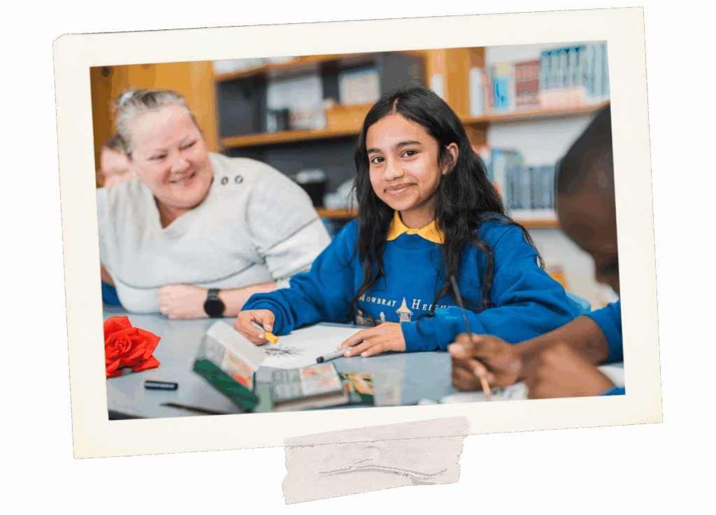 Young student smiling towards the camera (no teeth visible), they appear to using crayons. Teacher is out of focus but is smiling with teeth showing.